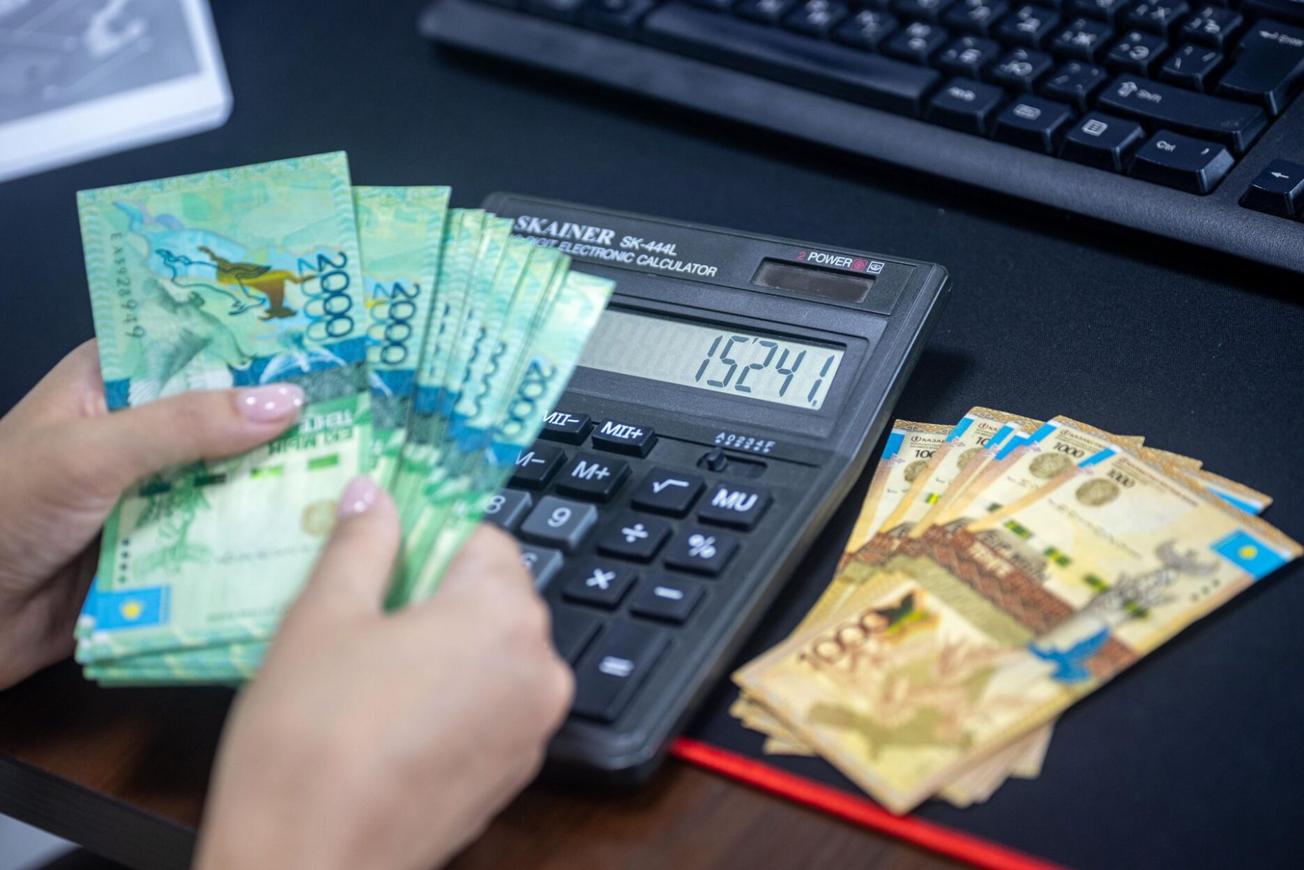 Рука пересчитывает купюры тенге рядом с калькулятором и компьютером