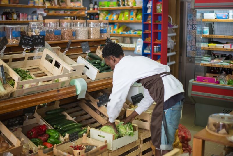 Работник продуктового магазина поправляет выкладку овощей на прилавке