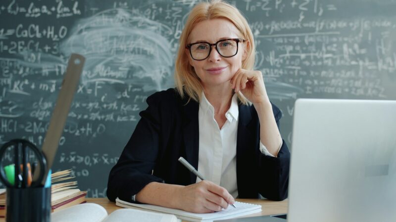 Учительница сидит за учительским столом в классе с ноутбуком и книгами